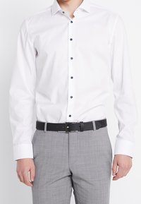 Vit långärmad skjorta med knappar i mörkblått, i kombination med ljusgrå texturerade byxor och ett svart bälte.