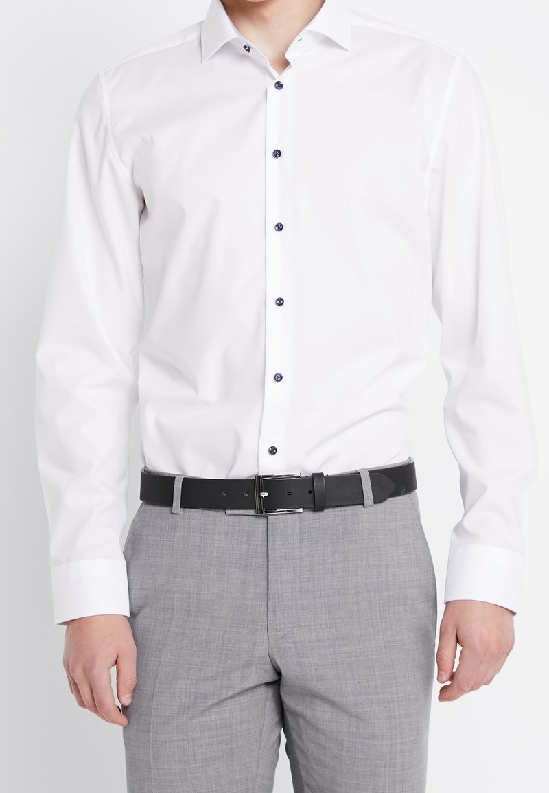 Vit långärmad skjorta med knappar i mörkblått, i kombination med ljusgrå texturerade byxor och ett svart bälte.