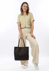 Černá kožená taška přes rameno s dvojitými uchy, obdélníkový tvar, hladká textura a minimalistický design. Model nosí světlý outfit.