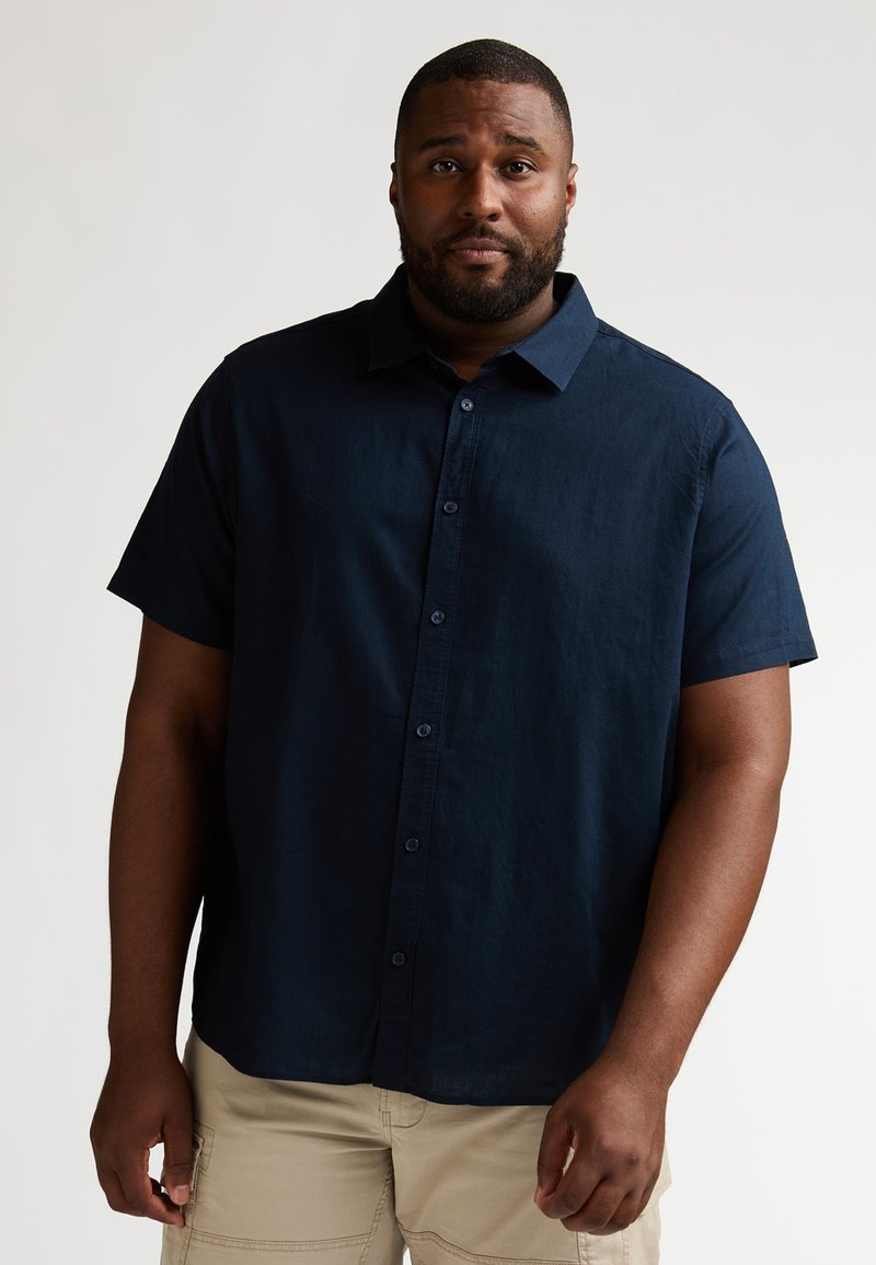 Chemise bleu marine à manches courtes avec boutons, fabriquée en tissu léger à texture lisse, dotée d'un col et d'une fermeture boutonnée sur le devant.