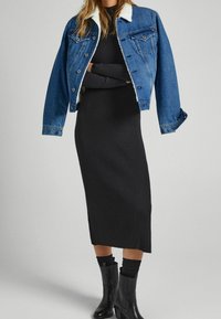 Džínová bunda s ovčí kožešinou na límci přes černé žebrované midi šaty. Modelka má na sobě černé kozačky po kolena s texturovaným detailen.