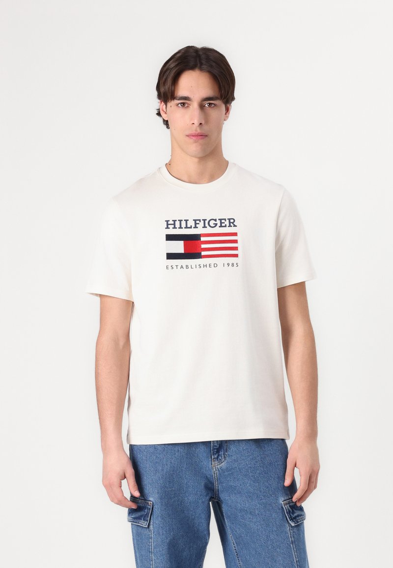 Krémové bavlněné tričko s krátkým rukávem, na kterém je grafické logo v námořnické modré a červené barvě, text "HILFIGER" a datum založení.