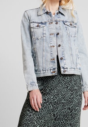 Kvinde iført en lyseblå denimjakke med kobberknapper og et grønt og sort leopardmønstret nederdel, foran en ensfarvet baggrund.