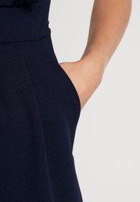 Námořnická modrá šaty s přiléhavým pasem a bočními kapsami, vyrobené z texturované tkaniny s hladkým povrchem. Viditelné detaily šití podél švu.