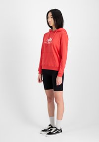 Sudadera roja con cordones y bolsillo frontal, con un logo blanco. Combinada con pantalones cortos de ciclismo negros y zapatillas negras con acentos blancos.