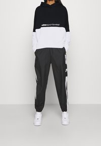 Henkilö, jolla on päällä musta ja valkoinen "nike sportswear" huppari, mustat urheiluhousut valkoisilla raidoilla ja valkoiset lenkkarit, seisomassa yksinkertaista taustaa vasten.