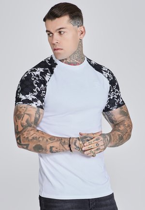 Tricou alb cu mâneci negre cu un model, având gât rotund și un design ajustat. Detalii notabile includ accente florale intricate pe mâneci.