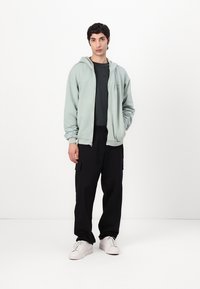 Felpa con zip verde chiaro, dal fit rilassato, indossata sopra una maglietta nera. Abbinata a pantaloni cargo neri e sneakers bianche. Texture morbida.