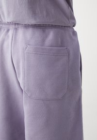 Gros plan d'une personne portant un pantalon de survêtement violet clair avec une poche arrière visible et un haut assorti sur un fond uni.