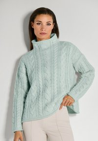 Frau trägt einen hellblauen grob gestrickten Rollkragenpullover und beigefarbene Hose, posiert mit einer Hand an der Hüfte vor einer weißen Wand.