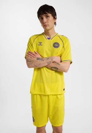Person med tatoveringer iført gul dansk fodboldtrøje og shorts, med armene over kors, stående foran en ensfarvet hvid baggrund.