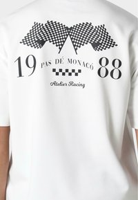Balta trumpų rankovių marškinėliai su juodu grafiniu dizainu, kuriame pavaizduoti šachmatų vėliavėlės, skaičiai 19 ir 88, taip pat tekstas "PAS DE MONACO" ir "Atelier Racing."