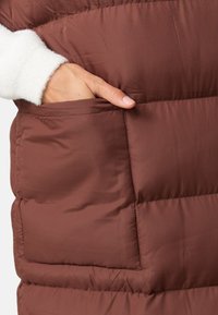 Ruskea paksu liivi, joka on valmistettu tikatusta kankaasta, sisältää sivutaskun ja valkoiset fleece-yksityiskohdat hihassa. Sileä pinta, lämmin muotoilu.