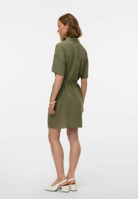 Abito a maniche corte di color verde oliva con colletto, vita cintata e vestibilità ampia, abbinato a sandali bianchi con tacco. Tessuto dalla texture liscia.