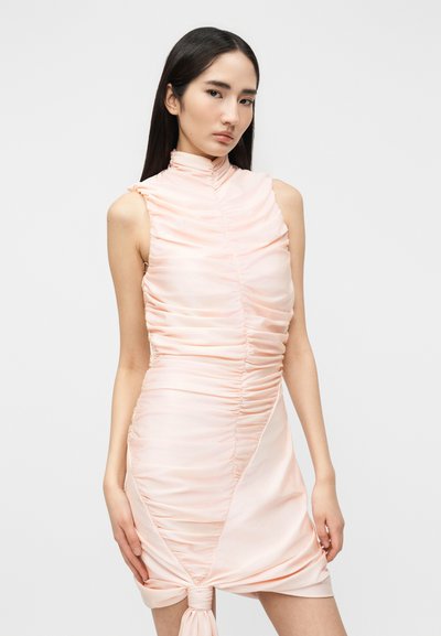 Νεαρή γυναίκα με μακριά σκούρα μαλλιά, που φοράει ένα αμάνικο ανοιχτό ροζ φόρεμα με ρυτιδώσεις και δεμένη λεπτομέρεια κόμπου στο τελείωμα, μπροστά σε ένα απλό φόντο.