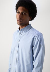 Lyseblå rutete button-up skjorte med en strukturert krage, lange ermer og hvite knapper. Stoffet virker glatt og godt tilpasset.