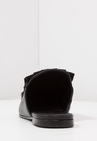 Svart sandal med öppen häl, platt sula och mjukt läderöverdrag. Har fransdetaljer längs sidorna för extra textur.