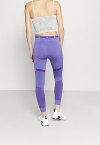 Fialové sportovní legíny s texturovanými panely a logem na pásu, v kombinaci s světlounce šedým přiléhavým crop topem a bílými teniskami.