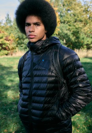 Schwarze Daunenjacke mit glänzender Textur, featuring einen hohen Kragen, Reißverschluss und Seitentaschen, getragen über einem grauen Hemd, vor grüner Kulisse.