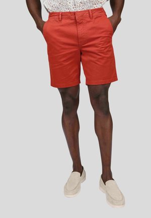 Shorts en coton rouges avec poches avant, face plate et coupe ajustée. Associés à des chaussures à enfiler de couleur claire.