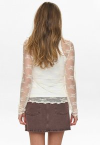 Witte langslijve top met kantdetails en bloemenpatroon, gecombineerd met een bruine denimrok. Kantafwerking aan de onderkant van de top.