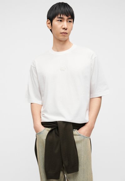 Λευκό βαμβακερό t-shirt με κοντά μανίκια και στρογγυλή λαιμόκοψη, με ένα διακριτικό κεντημένο λογότυπο στο στήθος· συνδυασμένο με ανοιχτόχρωμο ριγέ παντελόνι και σκούρο πράσινο πουλόβερ δεμένο γύρω από τη μέση.