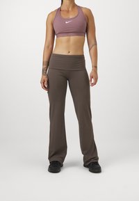 Fioletowy sportowy biustonosz z białym logo, w połączeniu z brązowymi rozkloszowanymi legginsami. Wykonane z elastycznego materiału, posiadają wysoki pas i gładką fakturę.