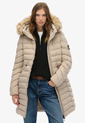 Beige pufferjas met een faux fur-gevoerde capuchon, rits aan de voorkant en geribbelde textuur. Draagt over een zwarte kabeltruien en blauwe jeans.