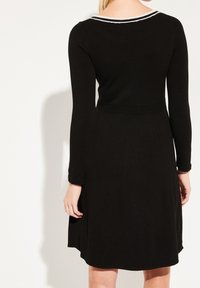 Robe noire à manches longues, avec un corsage ajusté et une jupe évasée. Présente un col rayé et une taille texturée. Tissu lisse.