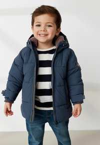 Marineblaues gefüttertes Jacket mit Kapuze aus Fleece, Reißverschluss vorne und Knopfakzenten. Getragen über einem gestreiften Shirt und kombiniert mit blauen Jeans.