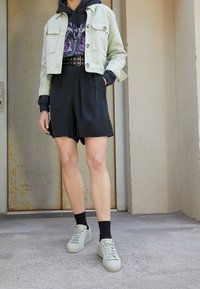 Jasnozielona skórzana kurtka typu cropped noszona na czarnej bluzie z kapturem, w połączeniu z czarnymi szortami i zielonymi sneakersami. Posiada teksturę materiału oraz detale z guzikami.