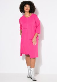 Abito rosa a maniche corte con due tasche frontali e orlo irregolare. Il modello indossa stivaletti alla caviglia argentati lucidi, mostrando un design casual ma moderno.