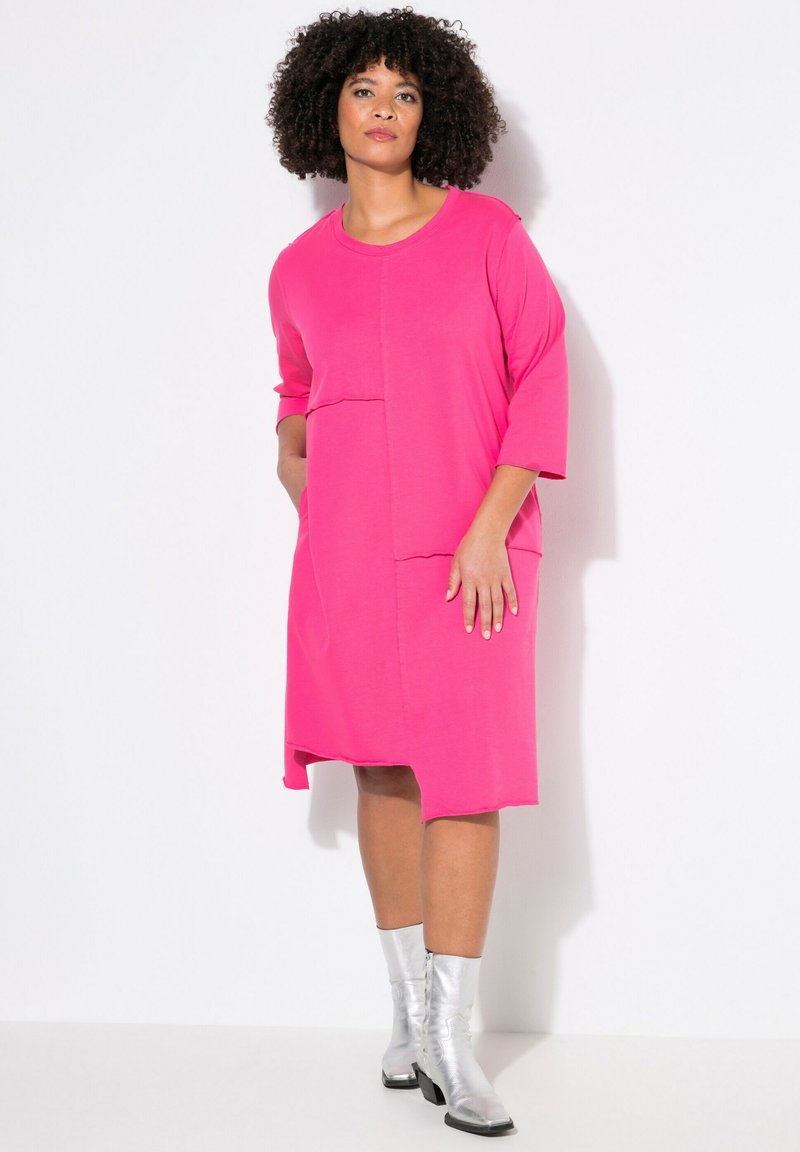 Abito rosa a maniche corte con due tasche frontali e orlo irregolare. Il modello indossa stivaletti alla caviglia argentati lucidi, mostrando un design casual ma moderno.