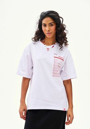 Kobieta z kręconymi włosami, ubrana w białą, oversize'ową koszulkę z czerwonym napisem i kolorowy naszyjnik z koralików, stojąca na tle jednolitego jasnego tła.