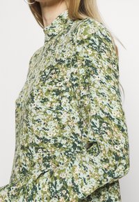 Hochgeschlossenes, langärmliges Oberteil mit grünem Blumenmuster in verschiedenen Rosatönen und Blau. Hergestellt aus einem weichen Stoff, mit lässigem Schnitt und seitlicher Tasche.