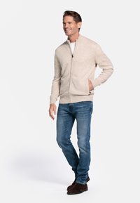 Beige ritsvest met geribbelde boorden en zoom. Draagt over een wit shirt, gecombineerd met blauwe jeans en donkerbruine schoenen.
