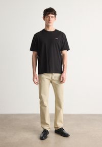 T-shirt en coton noir avec un petit logo sur la poitrine, associé à un jean beige clair et des chaussures noires. Design simple et coupe décontractée.