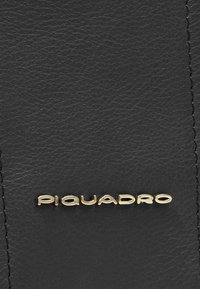 Schwarze Lederoberfläche mit einem goldenen "Piquadro"-Logo. Die Textur wirkt glatt mit sichtbaren Nähten entlang der Ränder.