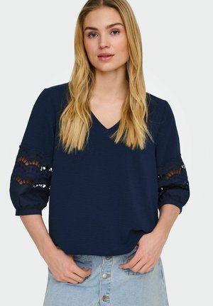 Lyshåret kvinde iført marineblå V-halsbluse med blonder på ærmerne og lyseblå knappede jeans, stående med hænderne nær lommerne.