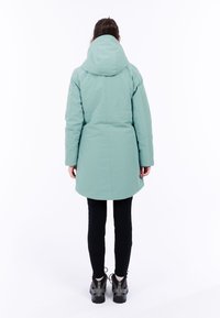 Mintgrön parka med huva, lätt avsmalnande design, sidofickor och längre bakstycke, matchad med svarta leggings och stövlar.