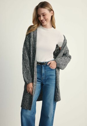Szary, dzianinowy kardigan z teksturowanym wzorem, otwarty przód, długie rękawy, noszony na dopasowanej białej bluzce i zestawiony z niebieskimi, szerokimi dżinsami.