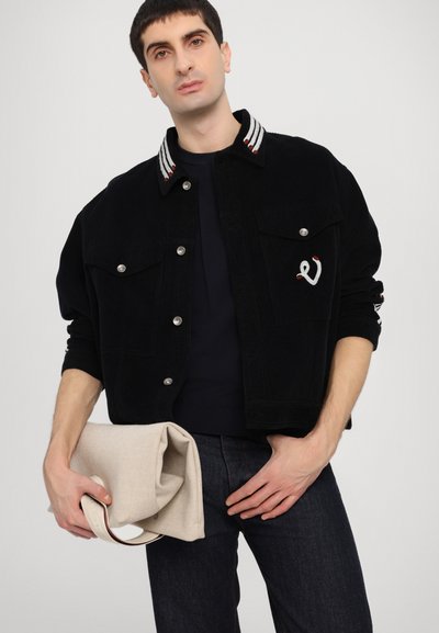 Giacca di velluto a coste nera con bottoni argentati, colletto a righe rosso e bianco, dettagli delle tasche. Borsa beige con tracolla tenuta in mano.
