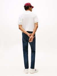 Bílá polo tričko, tmavě modré džíny a bílé tenisky. Muž s kudrnatými vlasy, který má na sobě vínovou čepici, s rukama sepnutýma za zády.