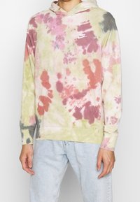 Mikina s kapucí ve stylu tie-dye s kombinací růžové, zelené a šedé barvy, s přední kapsou a malým bílým logem Nike na hrudi.