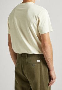 Kermakotoinen t-paita lyhyillä hihoilla, yhdistettynä oliivinvihreisiin housuihin, joissa on takatasku ja brändilappu. Yksinkertainen, rento muotoilu.