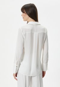 Witte blouse met lange mouwen van lichtgewicht, doorschijnende stof. Voorzien van een kraag, knoopmanchetten en een achterste plooi voor extra detail.