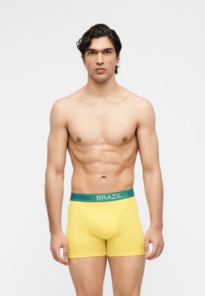 Tânăr cu păr închis la culoare, purtând chiloți boxer galbeni cu inscripțiile „BRAZIL” și „BJORN BORG” pe talie, stând în fața unui fundal simplu, deschis la culoare.