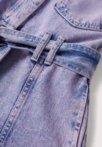 Abbigliamento in denim di colore lilla chiaro con accenti più scuri. Presenta una vita con cintura e tasche, tessuto testurizzato con schemi di sbiadimento.