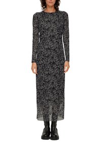 Langes schwarzes Kleid mit einem transparenten Überzug, das ein weißes abstraktes Muster aufweist. Lange Ärmel und ein gerader Schnitt bis zum Knöchel. Schwarze Knöchelstiefel.