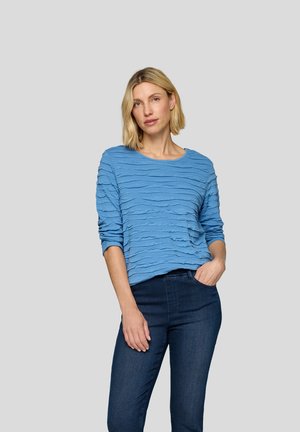 Helles blaues, strukturiertes Sweatshirt mit wellenförmigem Muster, runder Ausschnitt und langen Ärmeln, kombiniert mit dunklen Jeans. Modell steht vor einem grauen Hintergrund.